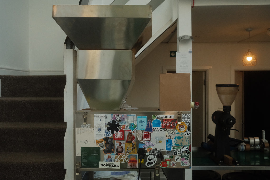A box of coffee concentrate placed on top of a stainless steel coffee bean hopper with a lower panel covered in colorful stickers