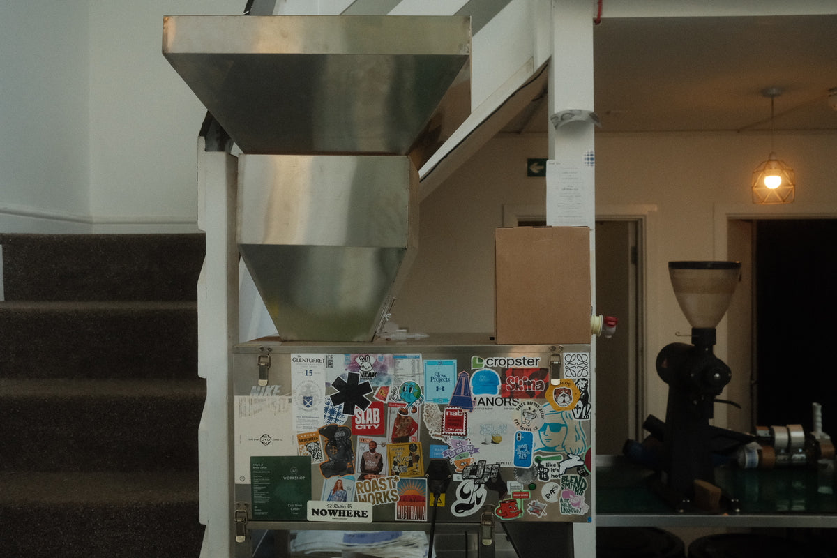 A box of coffee concentrate placed on top of a stainless steel coffee bean hopper with a lower panel covered in colorful stickers